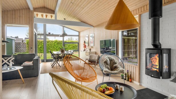 A cozy living room with a wooden ceiling, a black fireplace, and a large window overlooking a garden.