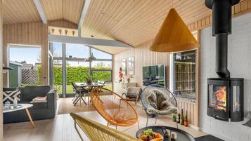 Un salon chaleureux avec un plafond en bois, une cheminée noire et une grande fenêtre donnant sur un jardin.