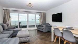 Ett modernt vardagsrum med ett stort fönster med utsikt över havet, en platt-TV som är monterad på väggen och ett matbord i trä med stolar.
