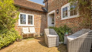 A brick house with a paved patio, two wicker sofas, a white bench, and a small garden area. - The Mews -  a beautiful cottage hidden away just off the village pond.  (Goudhurst)