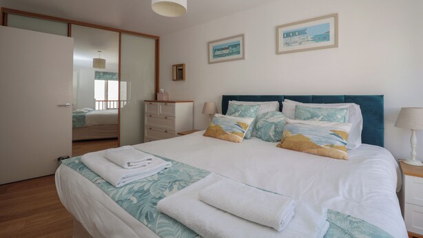 A neatly made bed with a blue and white floral bedspread, a teal headboard, and multiple pillows. There is a wooden dresser and a bedside table with a lamp. - Skylark (Reydon)
