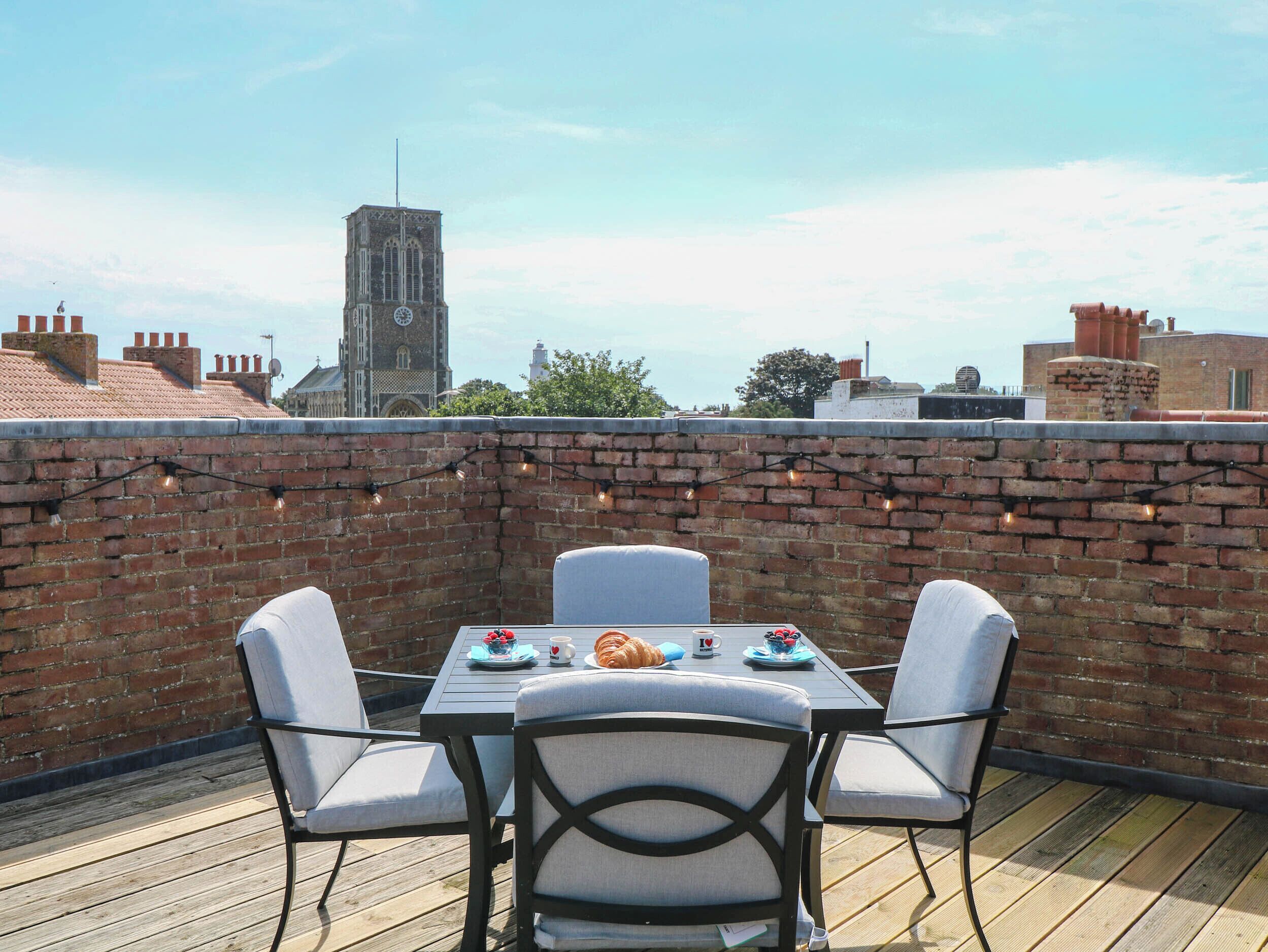 Una terraza en la azotea con un muro de ladrillo, una mesa para dos con sillas y una torre del reloj al fondo.