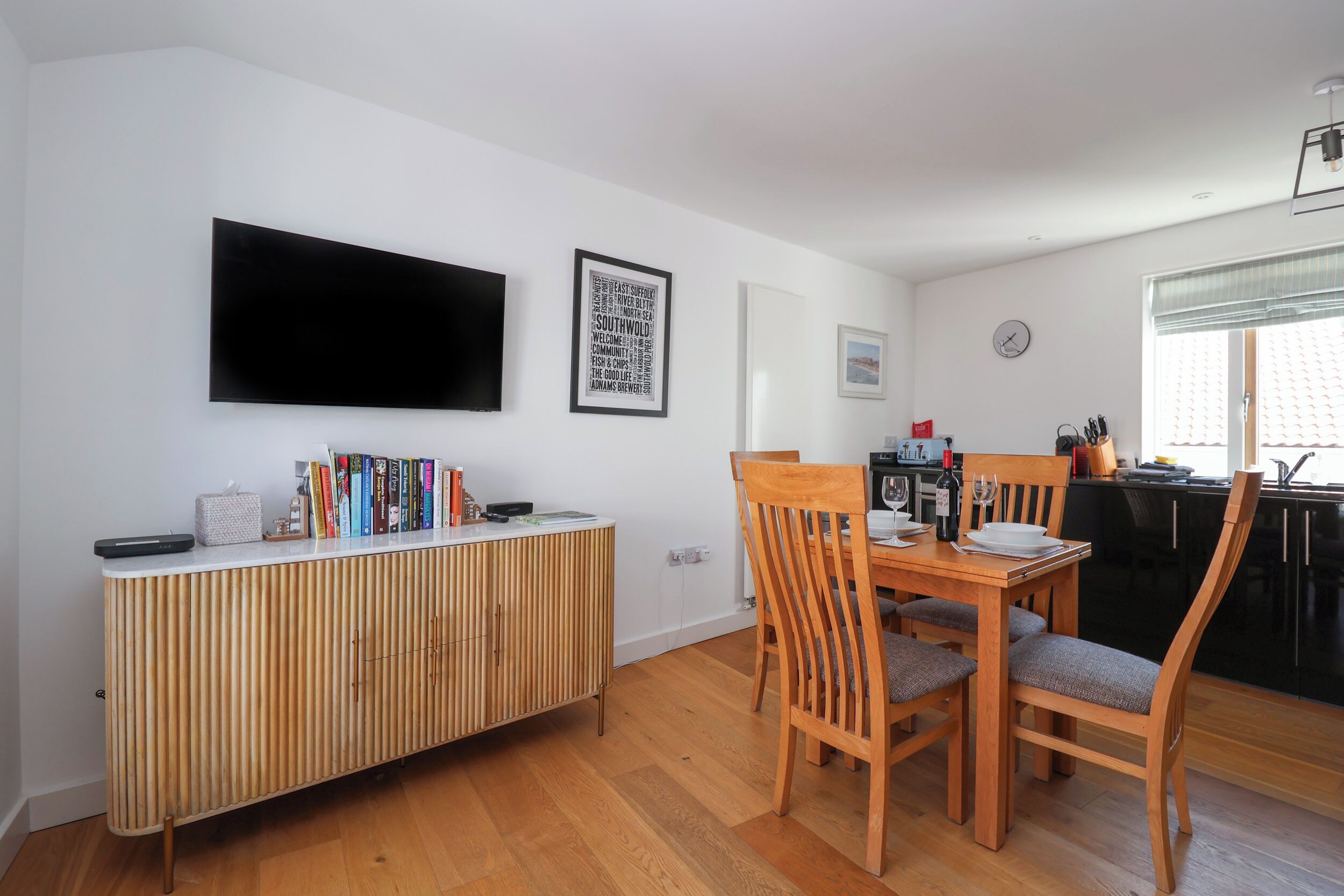 Una cocina moderna con comedor, que incluye una mesa de comedor de madera, sillas, un armario de madera empotrado, un televisor de pantalla plana en la pared y una ventana con vistas al exterior.