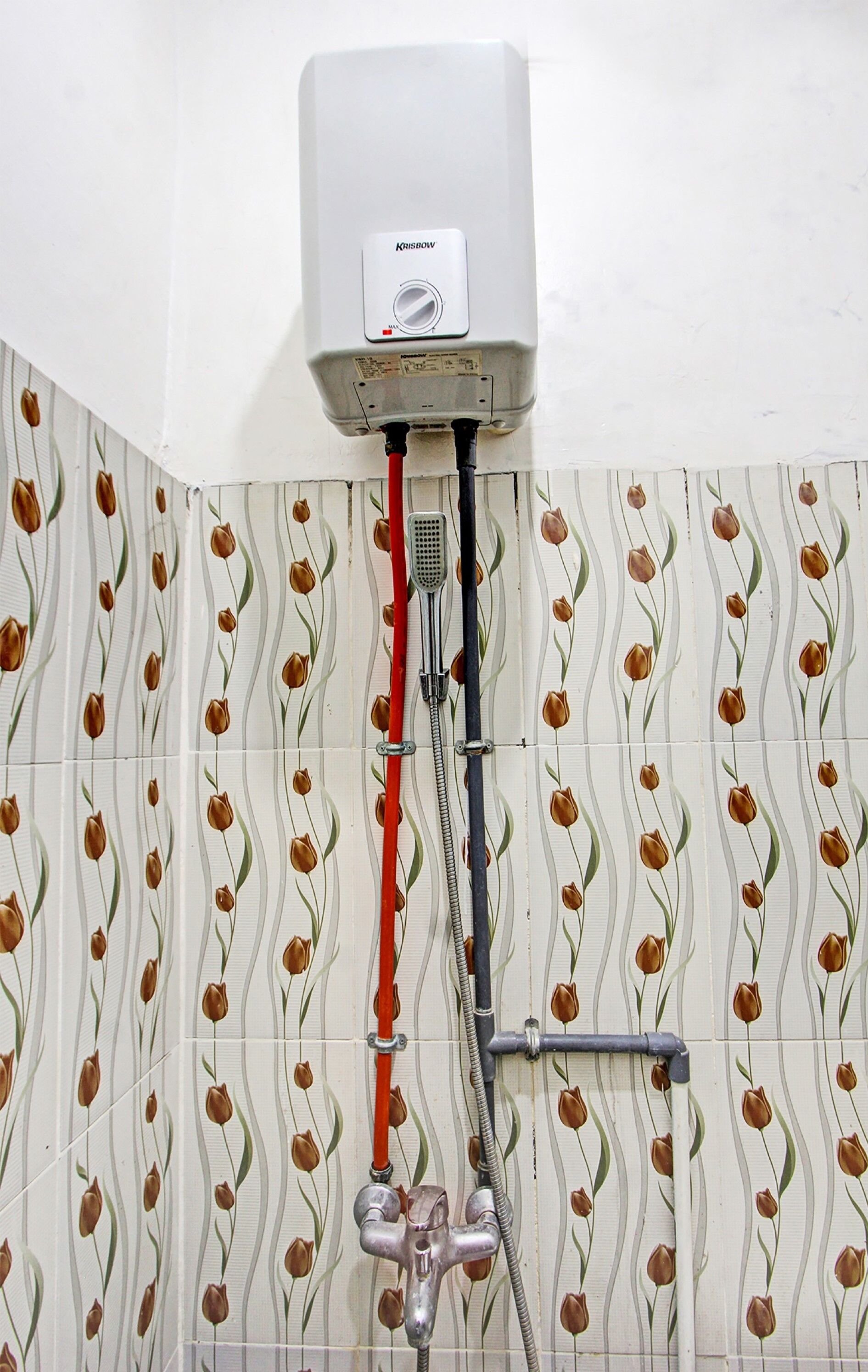 A bathroom with a white tiled wall featuring a tulip pattern, a Krissow water heater mounted above the wall, and a showerhead with a red and black hose.