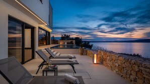 A modern outdoor patio with lounge chairs, a table with a bottle, and a stone wall overlooking a body of water during twilight. - Villa Ponti (Novigrad)
