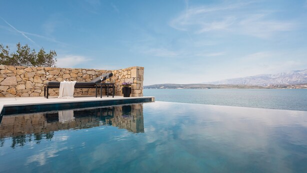 An infinity pool with a stone wall and seating area overlooking a body of water and distant mountains. - Villa Ponti (Novigrad)