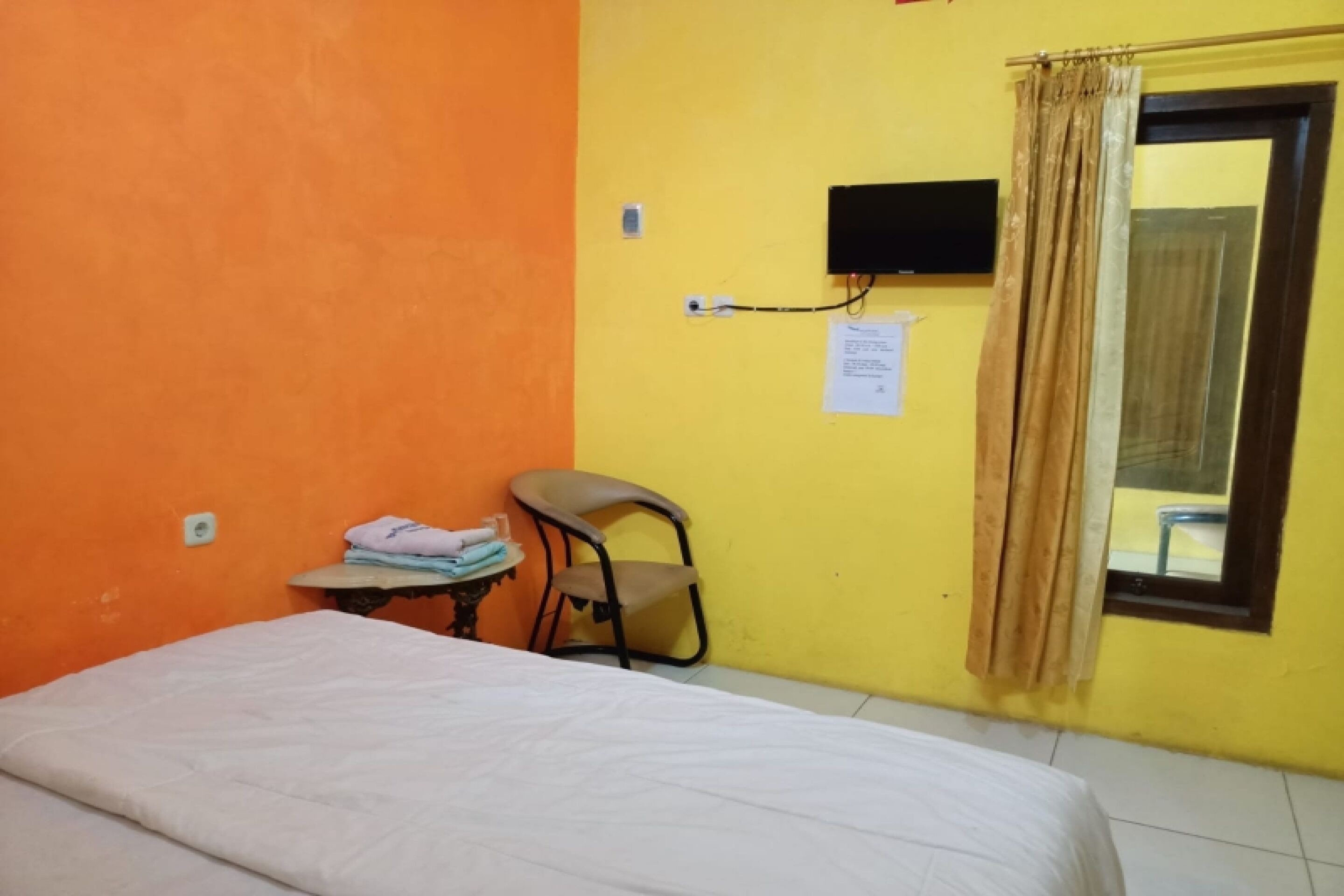 A room with a bed, a chair, a small table with towels, a wall-mounted TV, and a window with curtains.
