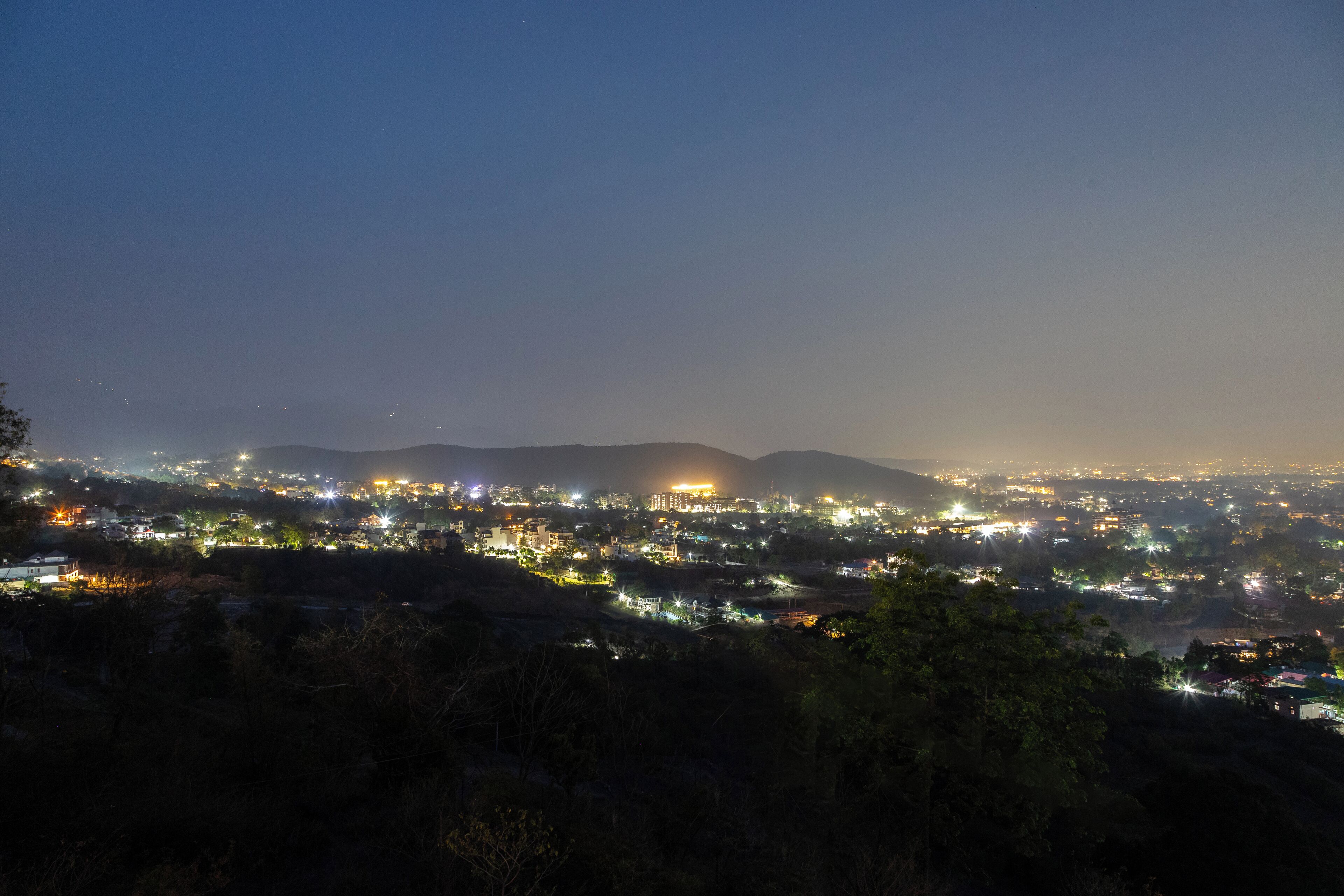 Une vue nocturne d’une ville, avec des bâtiments illuminés et des collines lointaines.