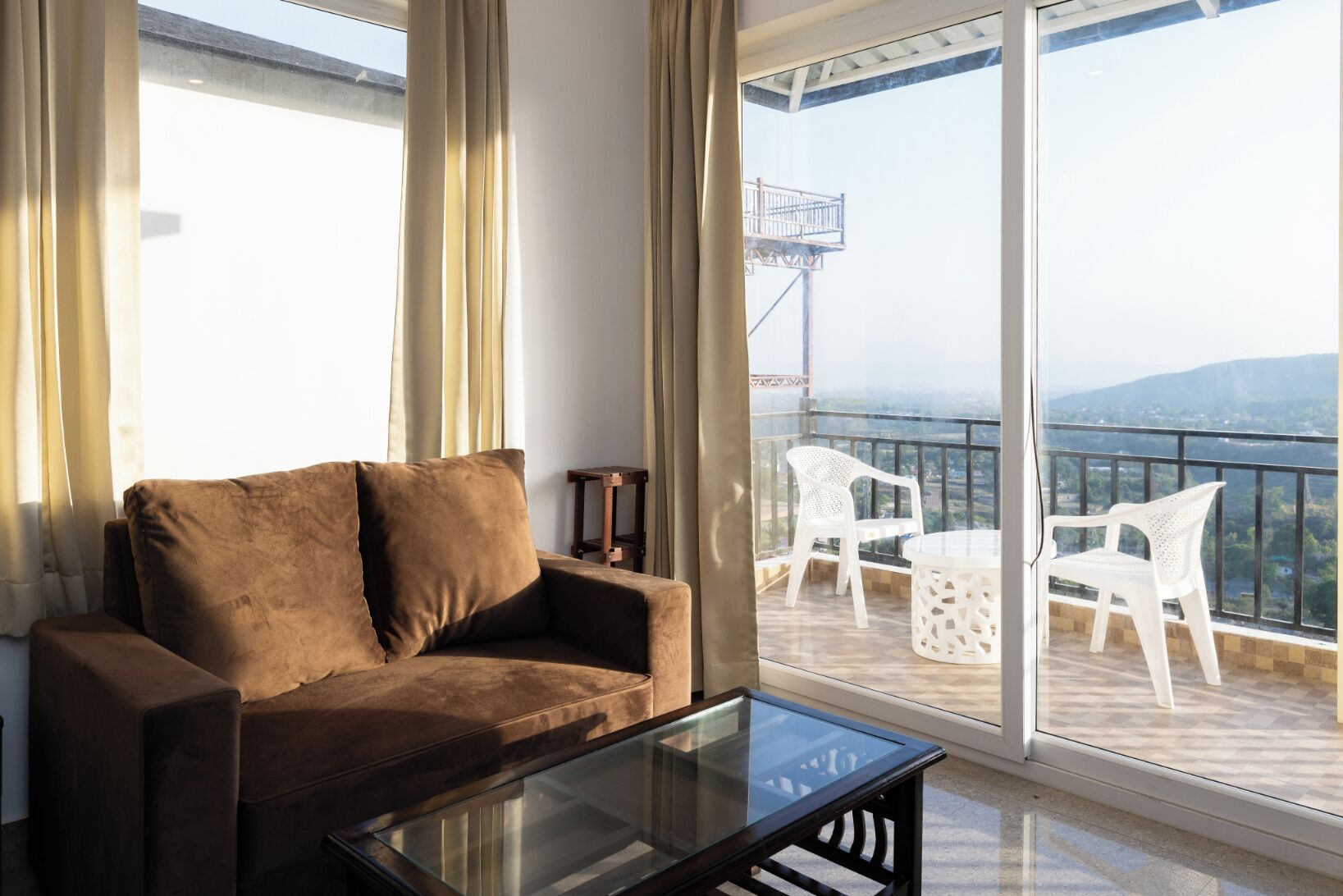 A room with a brown sofa, a glass coffee table, and a balcony with white chairs and a table.