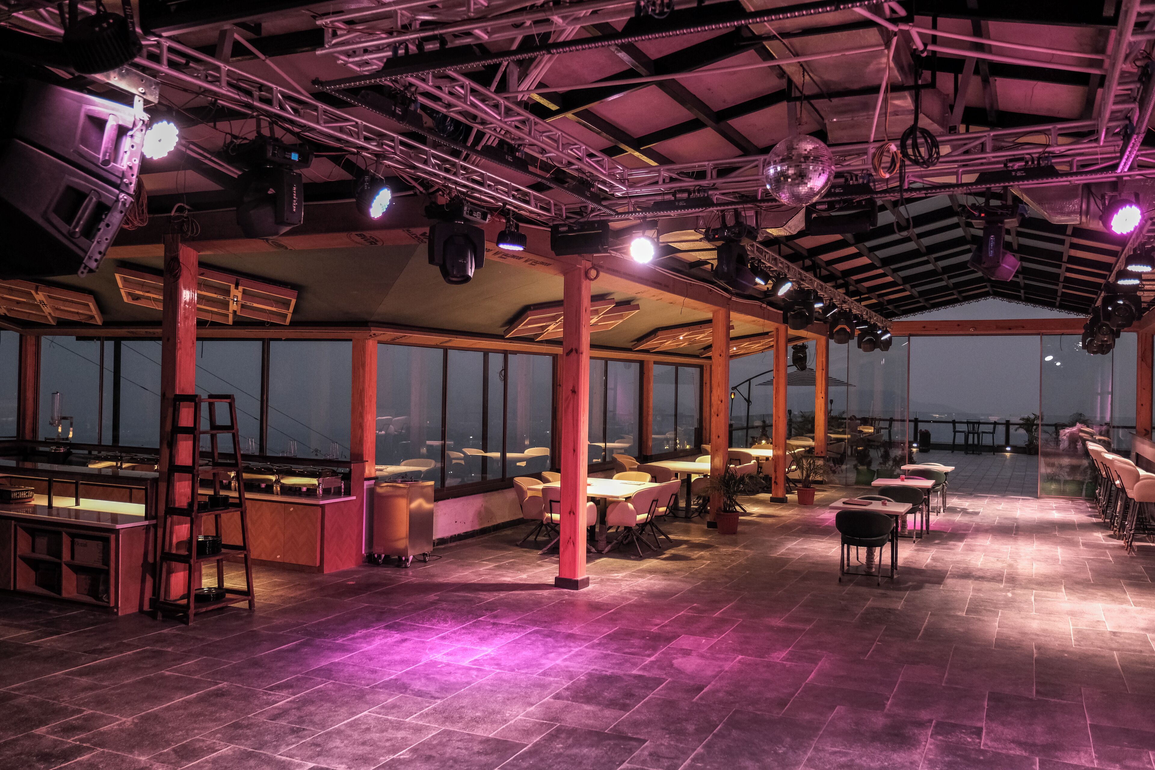 A spacious indoor area with tables and chairs, large windows, and a high ceiling with lighting fixtures.
