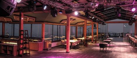 A spacious indoor area with tables and chairs, large windows, and a high ceiling with lighting fixtures.