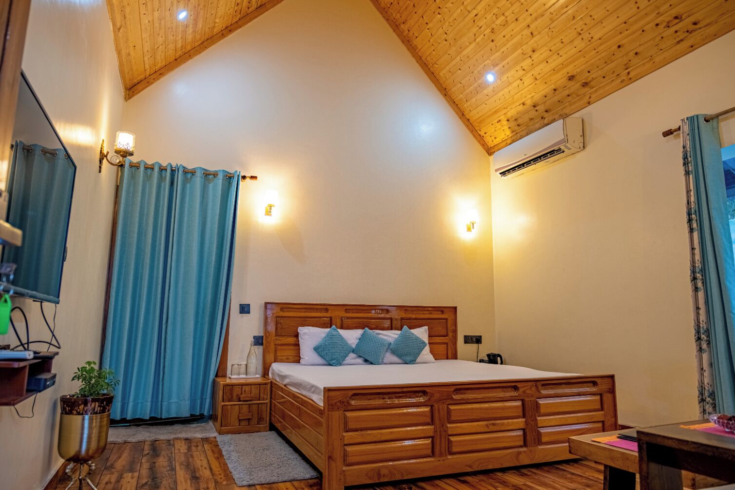 A bedroom with a wooden ceiling, a bed with a headboard, a bedside table, a lamp, a potted plant, and a window with curtains.