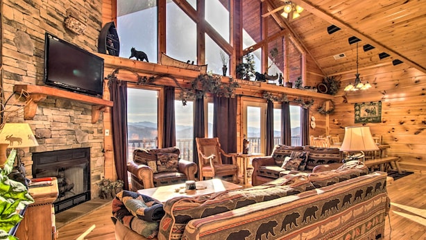 Interno di una baita in legno con camino in pietra, ampie finestre, televisore e divano con motivi animalier.