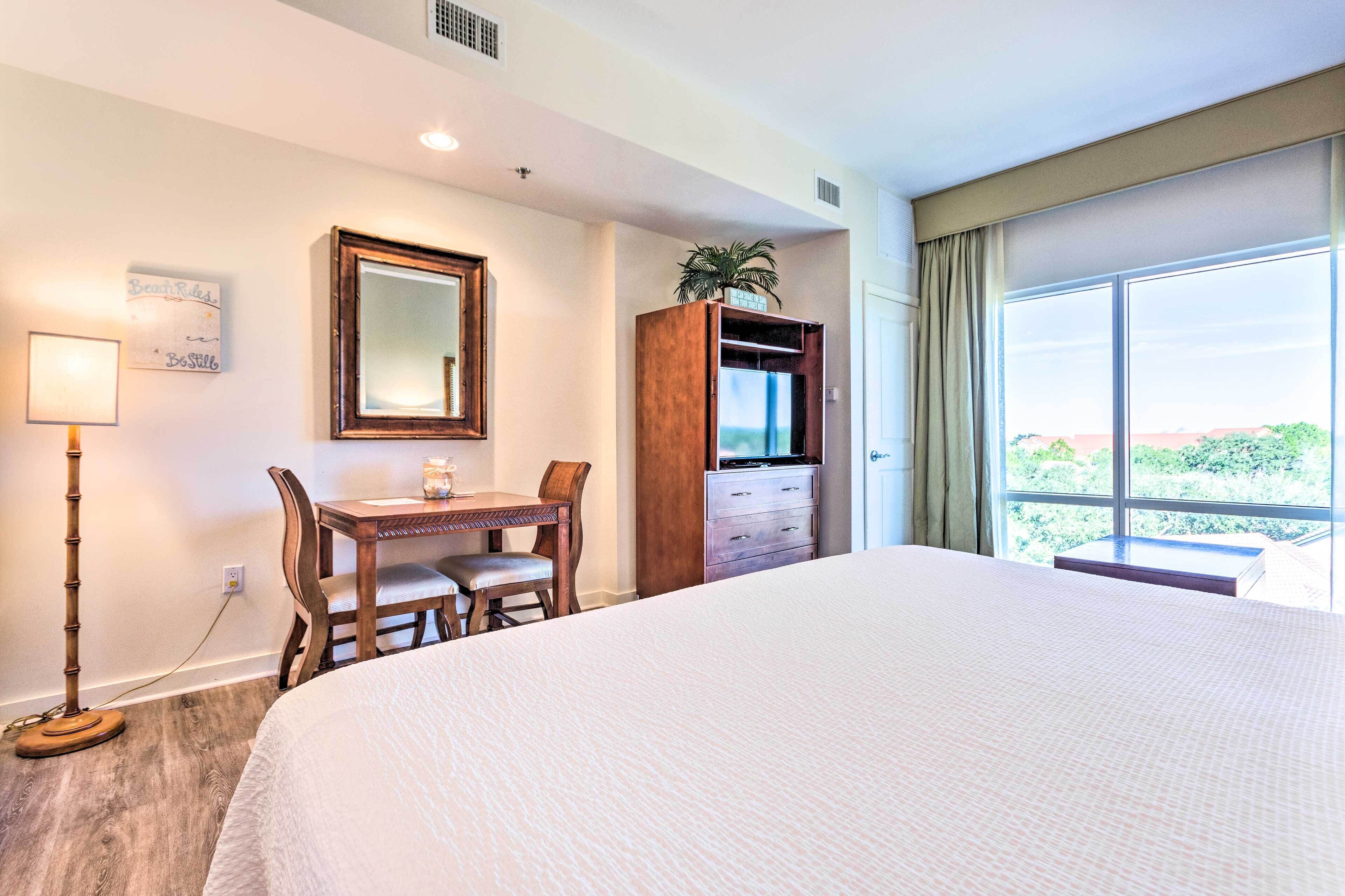 A hotel room with a large bed, a desk with a chair, a dresser, a mirror, and a window with a view.