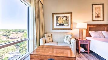 A hotel room with a large window, a sofa, a wooden chest, a bedside table with a lamp, and two framed pictures on the wall.