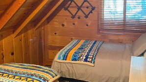 A cozy attic bedroom with a wooden sloped ceiling, a bed with patterned bedding, and a window with blinds. - Comfortable Mi-wuk Village Cabin: Hike & Ski! (Mi-Wuk Village)