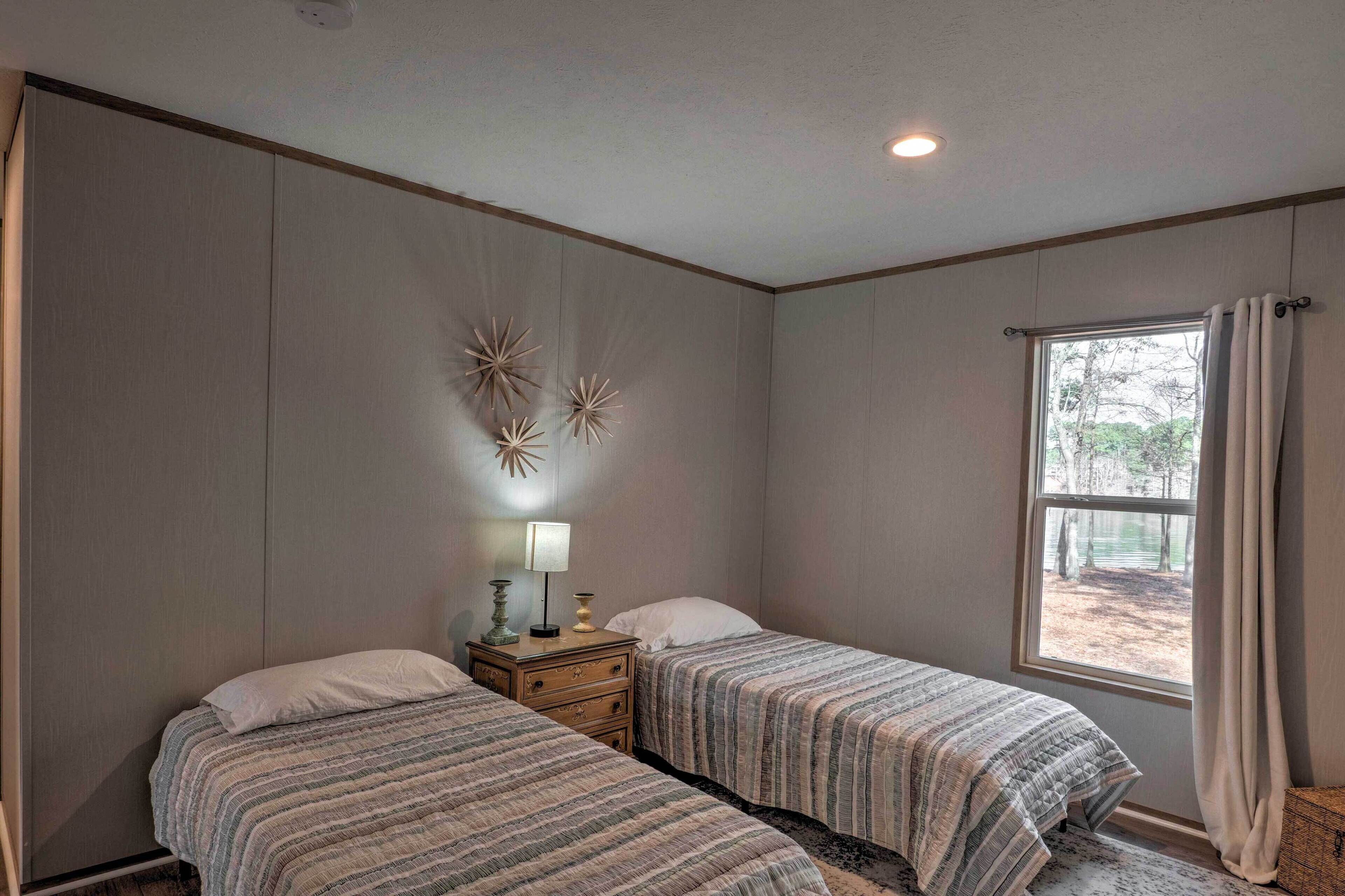 Une chambre avec deux lits, une table de chevet avec une lampe, des œuvres d’art décoratives au mur et une fenêtre avec des rideaux.