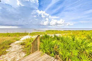 Un paseo marítimo de madera que conduce a una playa con vegetación verde y cielo despejado.