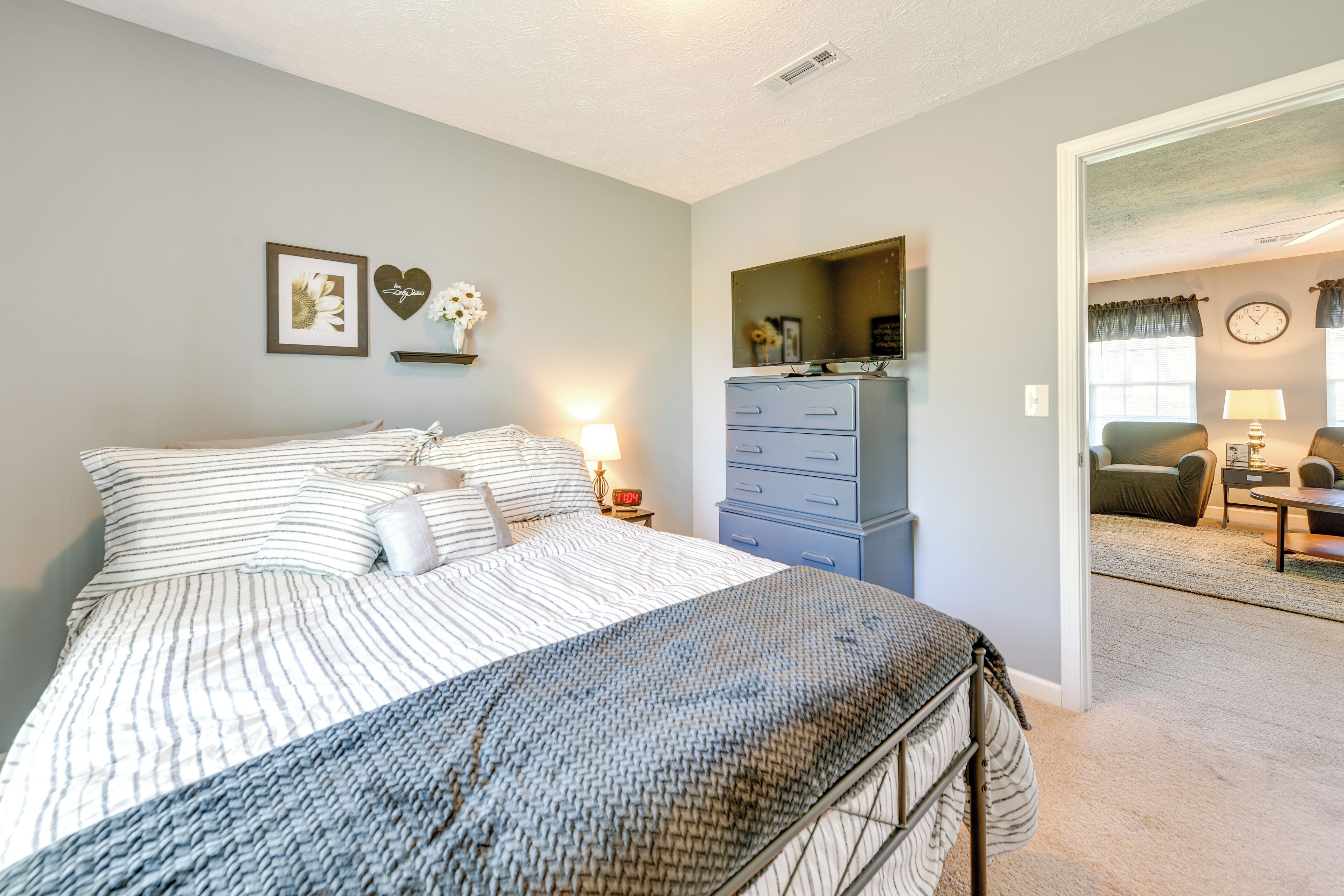 A bedroom with a bed, dresser, and a mirror. There is a door leading to another room with a chair and a table.