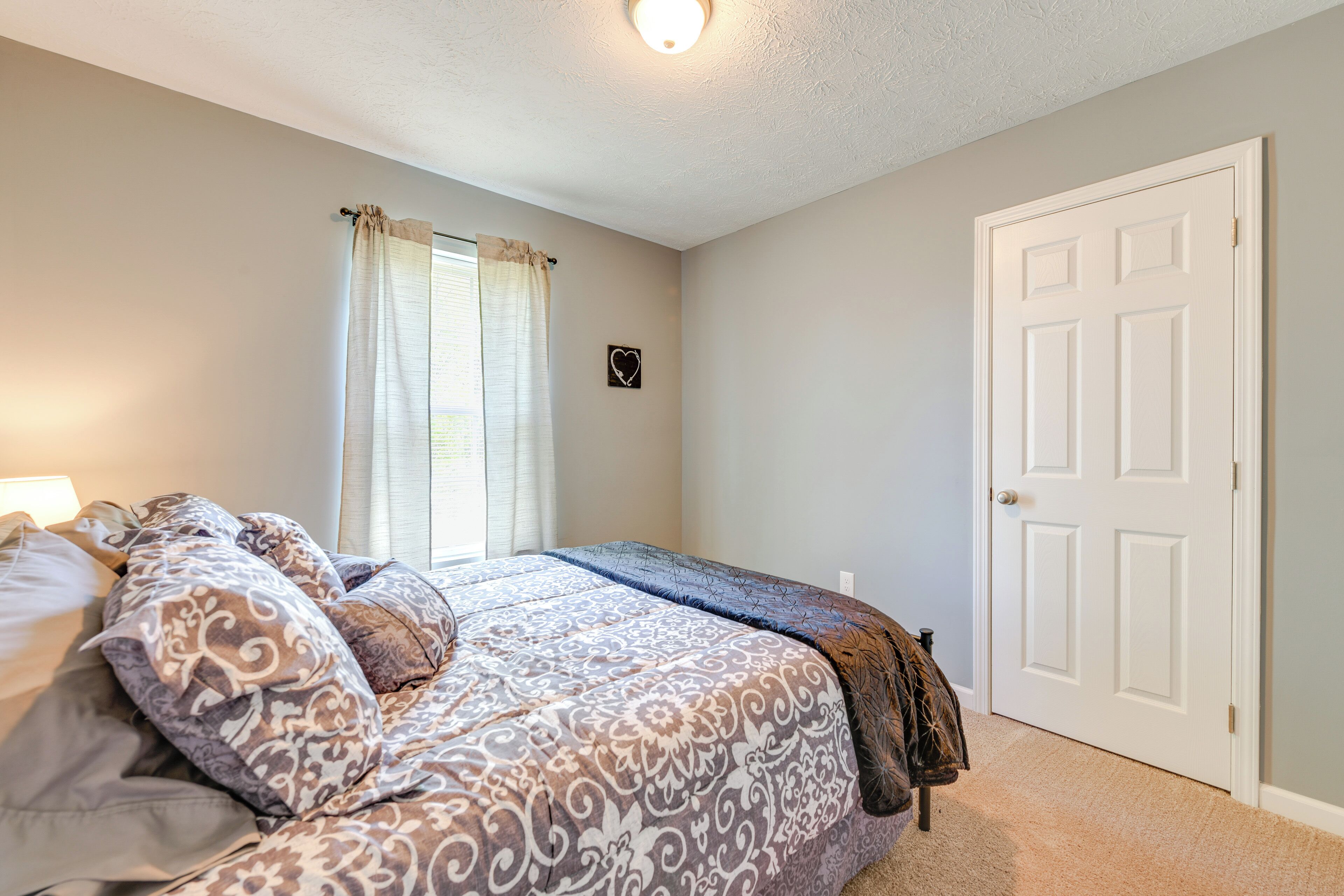 A bedroom with a bed, a window with curtains, a door, and a wall-mounted heart decoration.