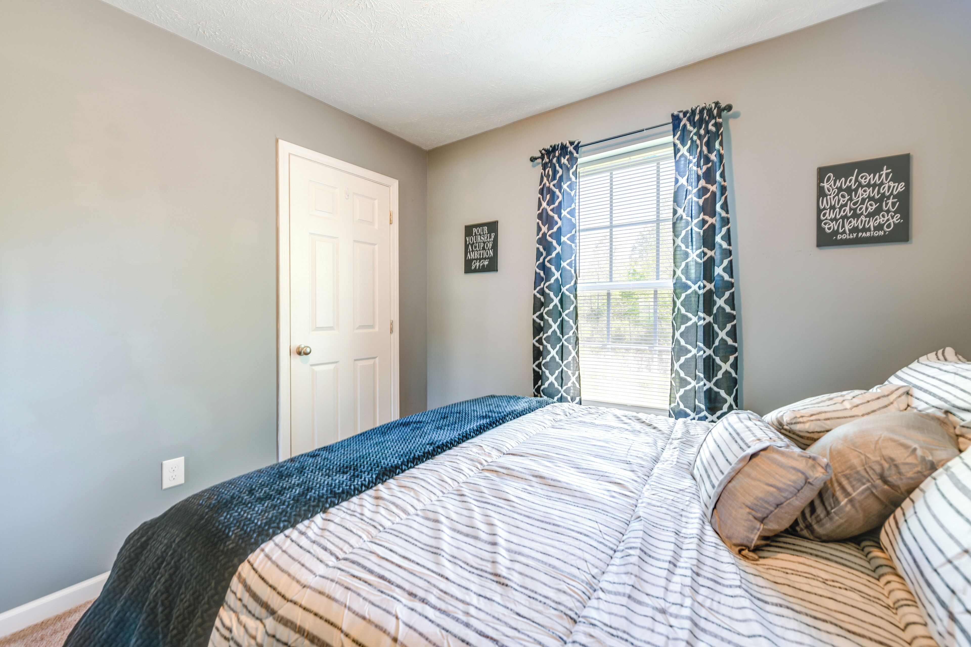 A bedroom with a bed, a window with patterned curtains, a door, and two wall decorations.