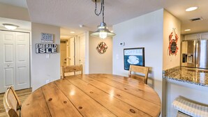 A dining area with a round wooden table, chairs, a hanging light fixture, and a kitchen with a stainless steel refrigerator. - Emerald Isle Condo w/ Direct Access to Ocean! (Emerald Isle)
