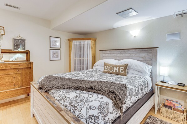 Quarto com cama de madeira, mesa de cabeceira, cômoda e janela com cortinas leves.