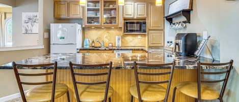 A kitchen with a central island, bar stools, wooden cabinets, a refrigerator, a microwave, and a coffee maker.