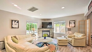 A spacious living room with a stone fireplace, beige sofas, a round coffee table, and large windows offering a view of greenery outside. - Family-friendly Kirkland Home ~ 1 Mi to Downtown (Kirkland)