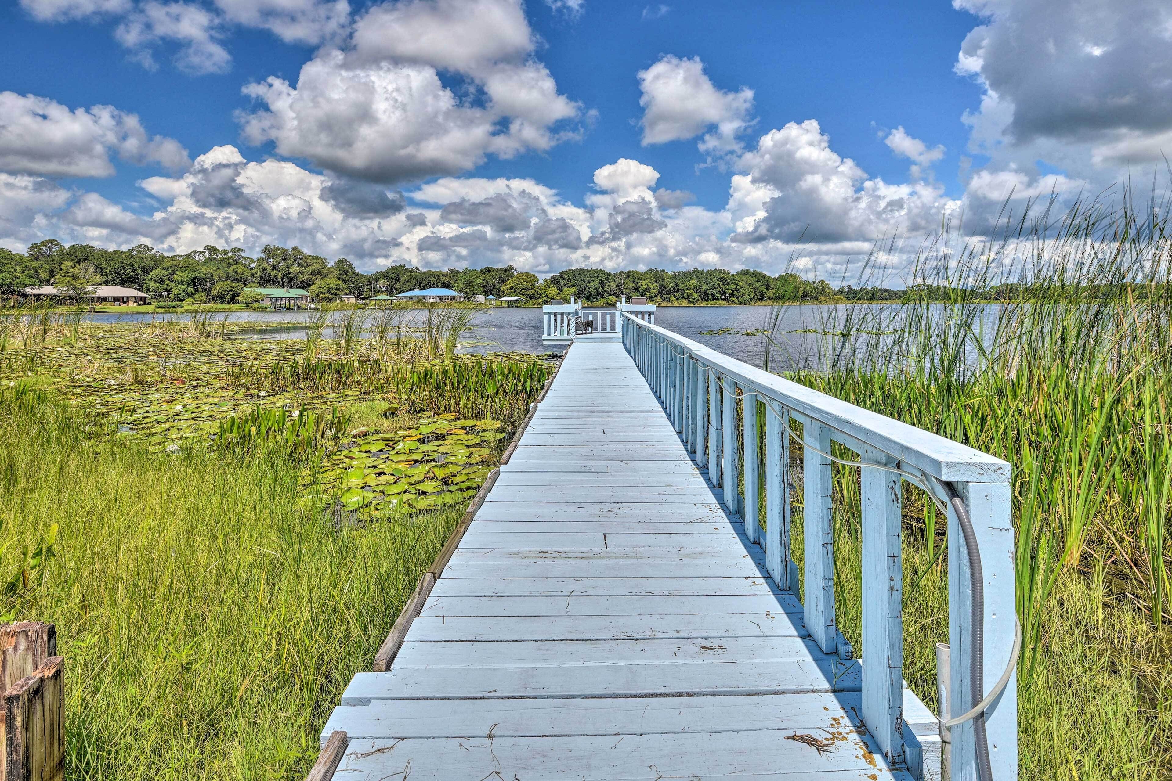 A wooden boardwalk extending over a body of water, surrounded by lush greenery and a clear sky.
