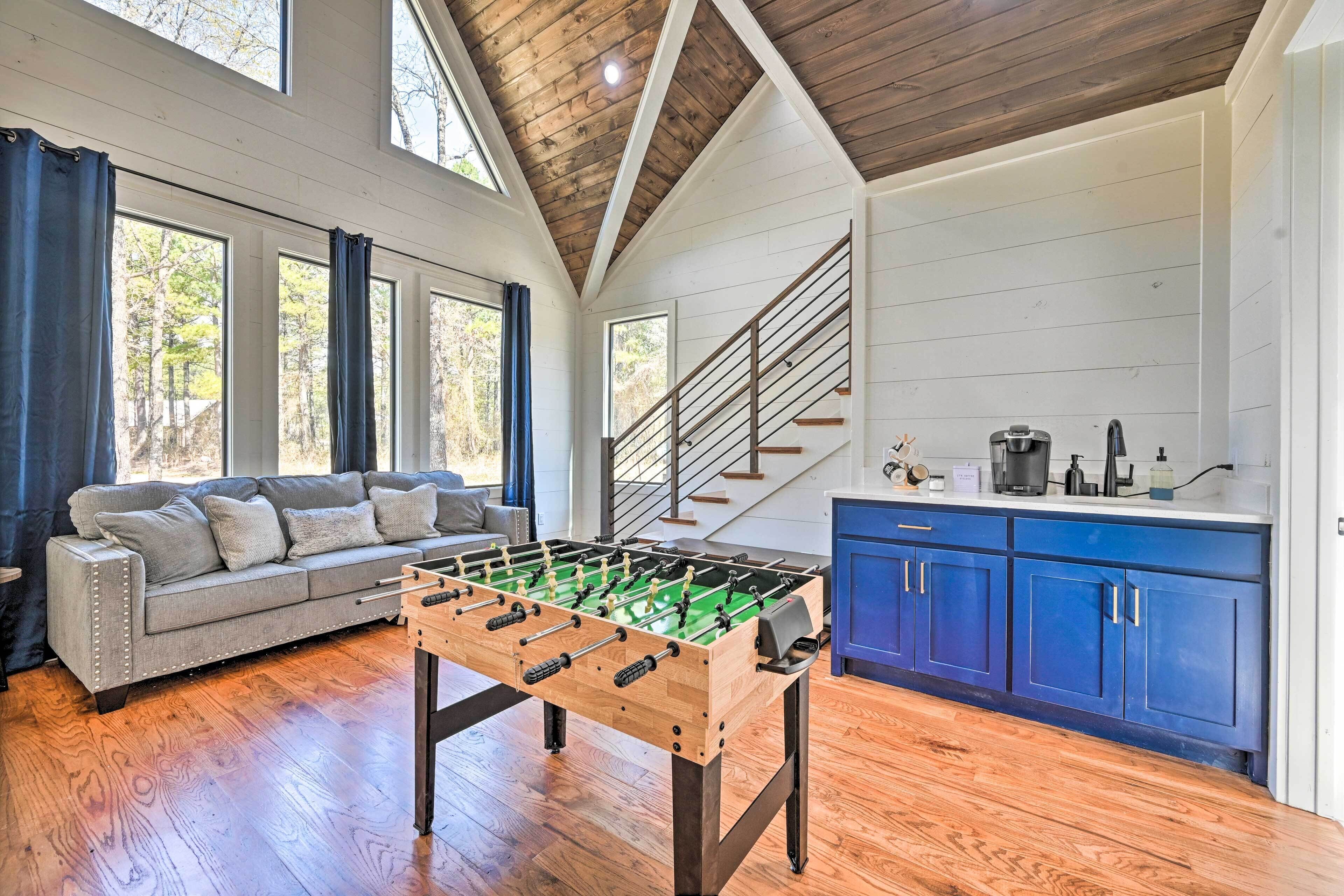 A spacious room with a wooden floor, a foosball table, a gray sofa, and a kitchen area with blue cabinets and a staircase.