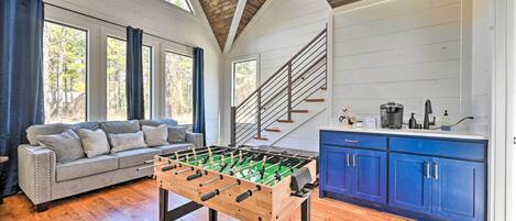 A spacious room with a wooden floor, a foosball table, a gray sofa, and a kitchen area with blue cabinets and a staircase.