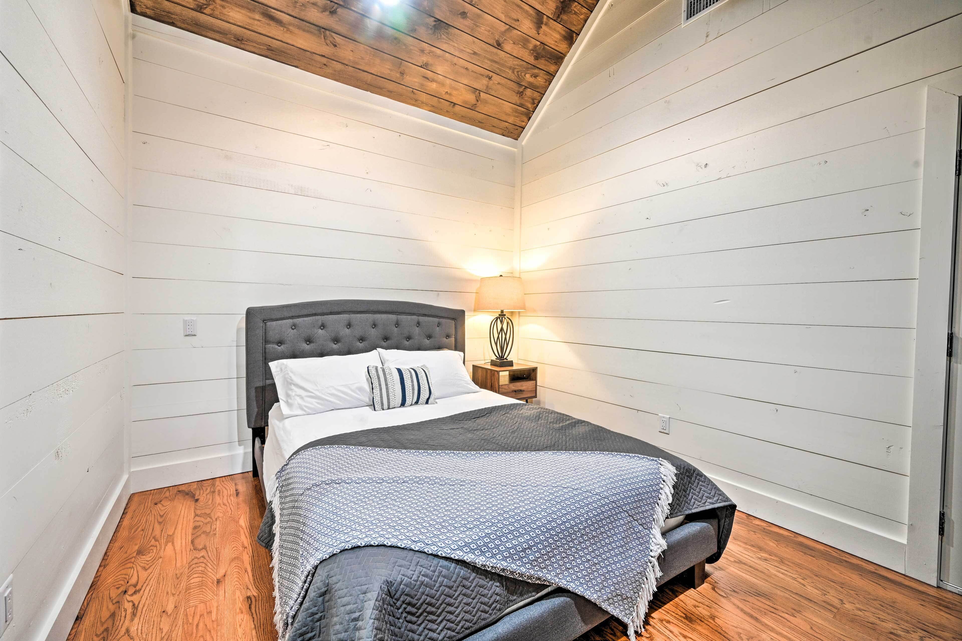 A bedroom with a bed, wooden flooring, a nightstand with a lamp, and a sloped ceiling.