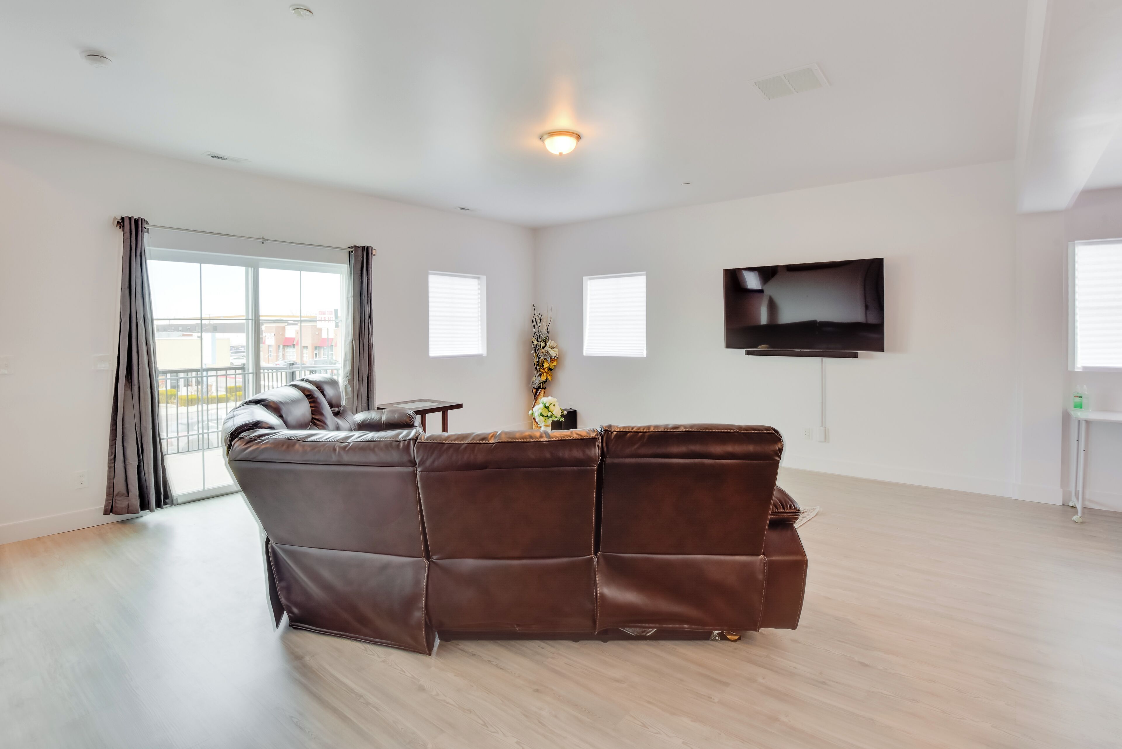 A spacious living room with a brown leather sectional sofa, a flat-screen TV mounted on the wall, and a large window with curtains.