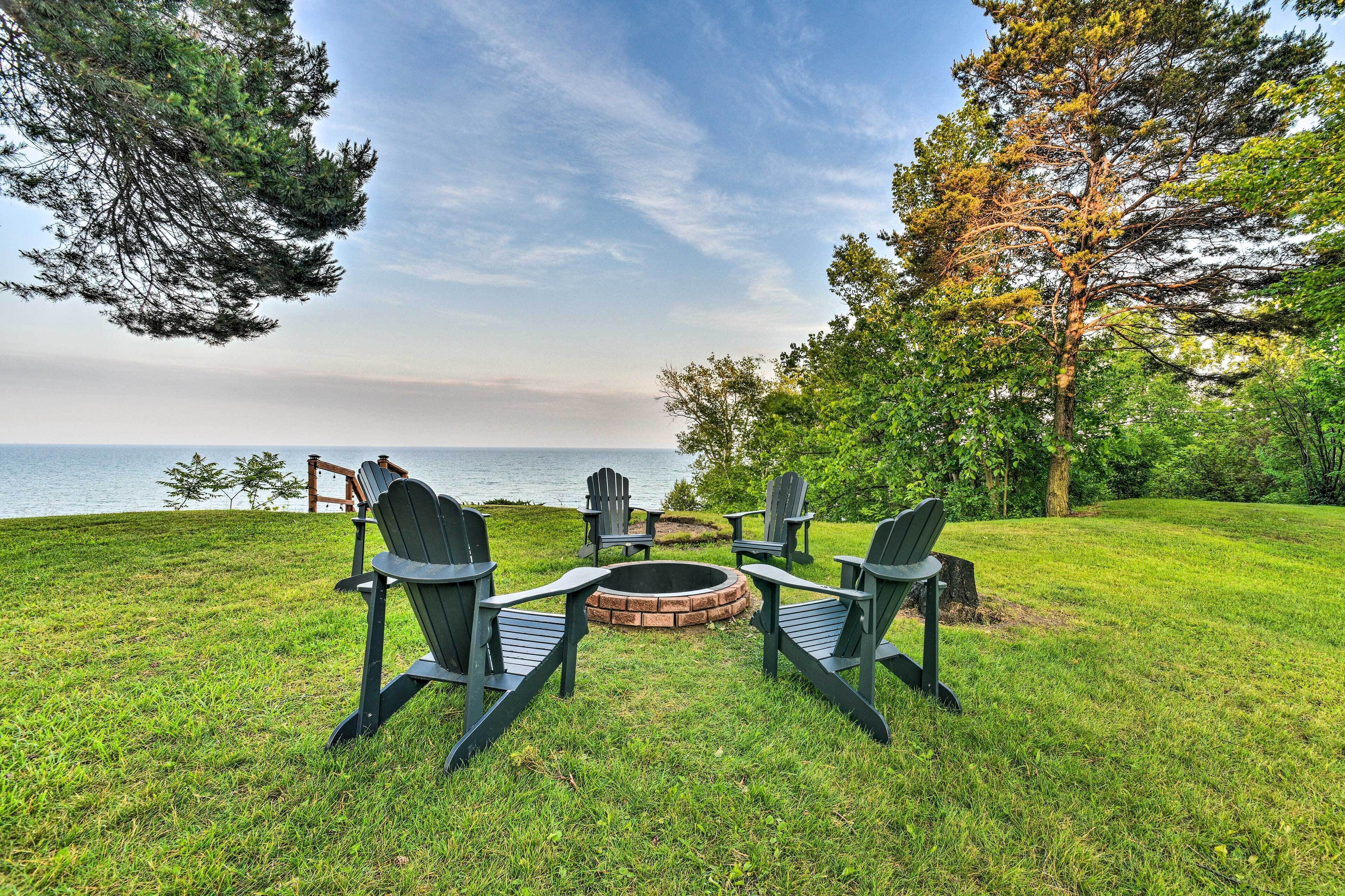 A grassy outdoor area with a fire pit and Adirondack chairs facing a body of water.