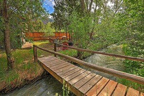 A wooden bridge over a stream with a wooden deck, surrounded by lush greenery and a wooden fence in the background.