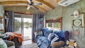 A cozy cabin interior with a wooden ceiling, a ceiling fan, a large window with curtains, a blue sofa, and a small table with a lamp. - Bay St Louis Houseboat 6 Mi to Old Town & Beach! (Bay St Louis)