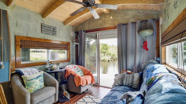 A cozy cabin interior with a ceiling fan, a large window with curtains, a sofa, a chair, and a small table with a lamp. - Bay St Louis Houseboat 6 Mi to Old Town & Beach! (Bay St Louis)