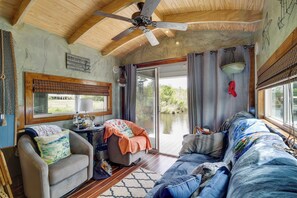 A cozy cabin interior with a ceiling fan, a large window with curtains, a sofa, a chair, and a small table with a lamp. - Bay St Louis Houseboat 6 Mi to Old Town & Beach! (Bay St Louis)