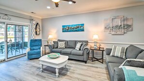 A living room with a grey sofa, blue armchair, and a white coffee table. There are two side tables with lamps. A painting of birds is on the wall, and a sign reads "Welcome to the Beach." - Screened Porch + Golf On-site: Myrtle Beach Condo (Myrtle Beach)