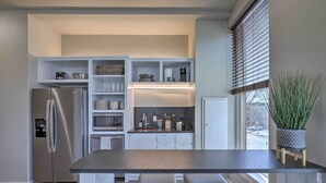 Une cuisine moderne avec des meubles blancs, un réfrigérateur en acier inoxydable, un four encastré et une grande fenêtre équipée de stores.