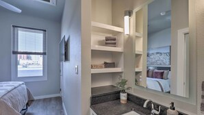 Une chambre d’hôtel avec un lit, une fenêtre avec des stores, une étagère intégrée avec des serviettes, un lavabo avec un miroir et une vue sur une montagne qui se reflète dans le miroir.