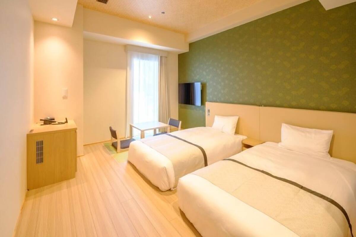 A hotel room with two beds, a small table, a chair, a desk, and a television on the wall.