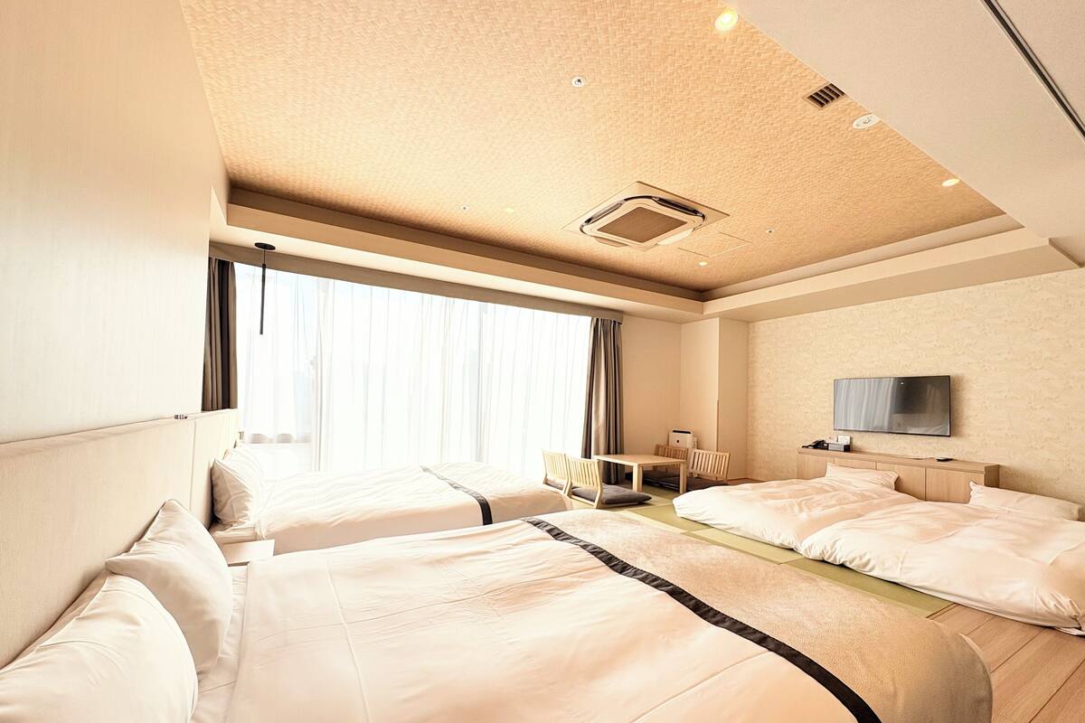 A hotel room with two beds, a desk, a TV, and a large window with curtains.