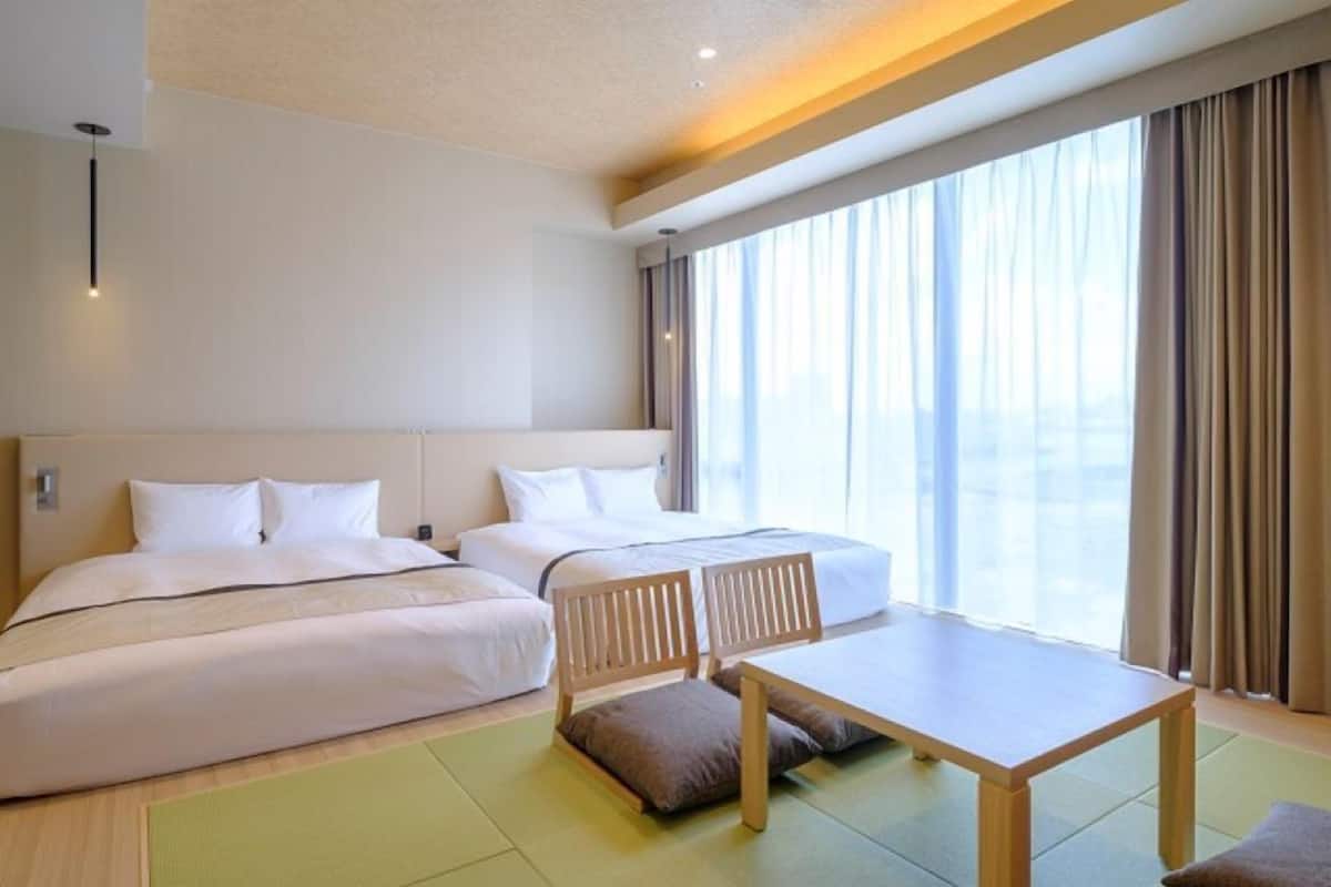 A hotel room with two beds, a dining table, and a view of the city through large windows.