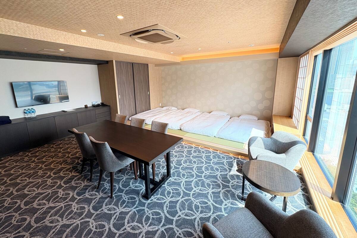A hotel room with a large bed, a dining table with chairs, a TV on the wall, and a view of the city through the window.