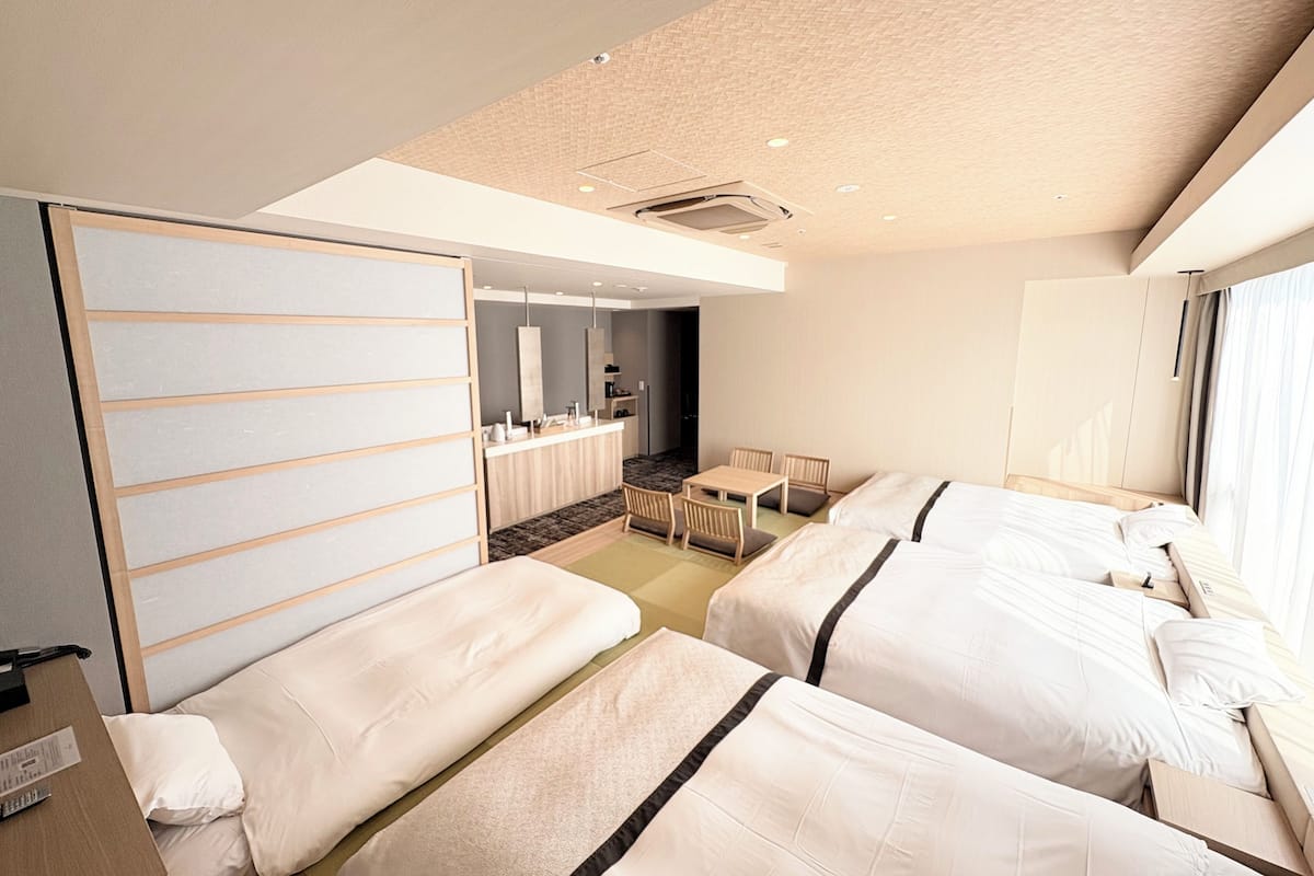 A hotel room with two beds, a small sitting area, a kitchenette, and a large window.