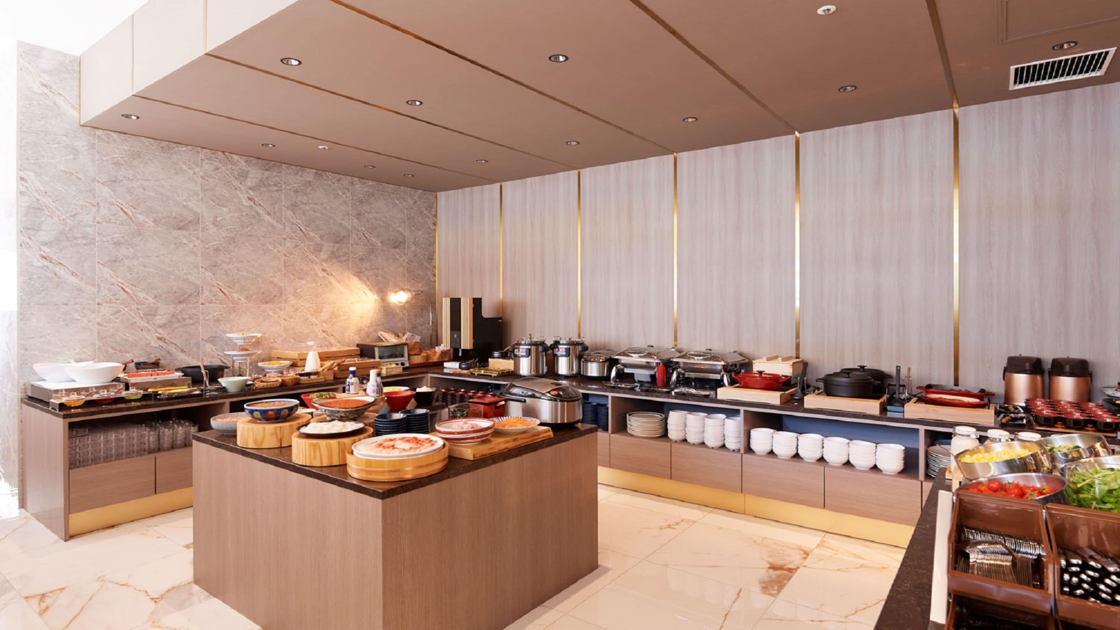 A modern buffet setup with a marble-tiled wall, wooden counters, and various food and drink containers.
