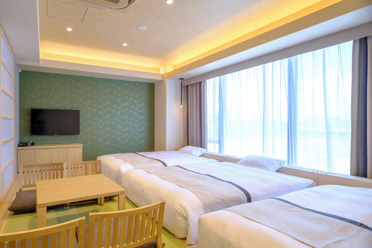 A hotel room with two beds, a TV, a dining table, and a view of the city.