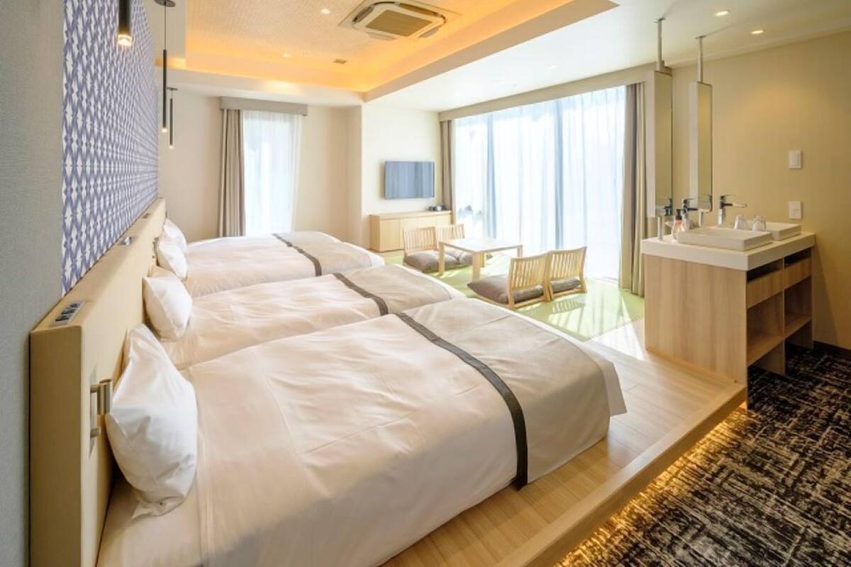 A hotel room with two beds, a desk, a chair, a large window, and a bathroom with a mirror and sink.